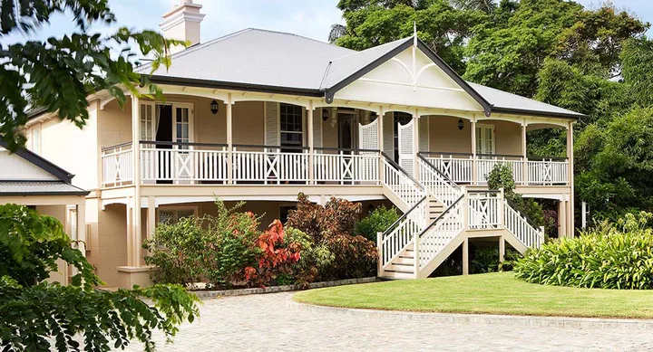 Elegant two-story house with wraparound balcony, surrounded by lush greenery and a manicured lawn.
