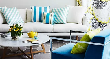 Living room with white sofa, striped cushions, blue armchair, marble coffee table with magazines, plants, and a yellow teapot.