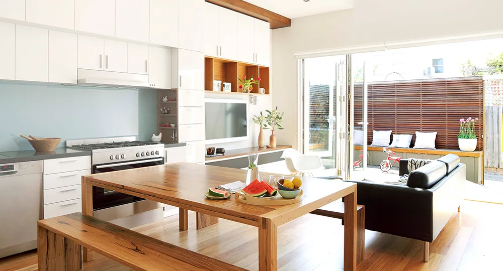 Bright modern kitchen-living space with wooden table, fruit bowl, black sofa, and glass doors opening to patio.