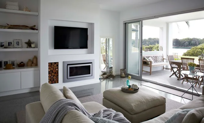 Stylish modern living room with beige sofa, TV, fireplace, and open sliding doors to a patio overlooking greenery and water.