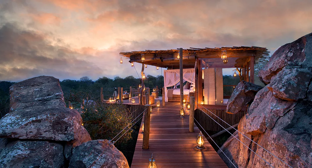 Elevated wooden deck with canopy bed surrounded by lanterns, set against a dramatic sunset sky among large rocks.