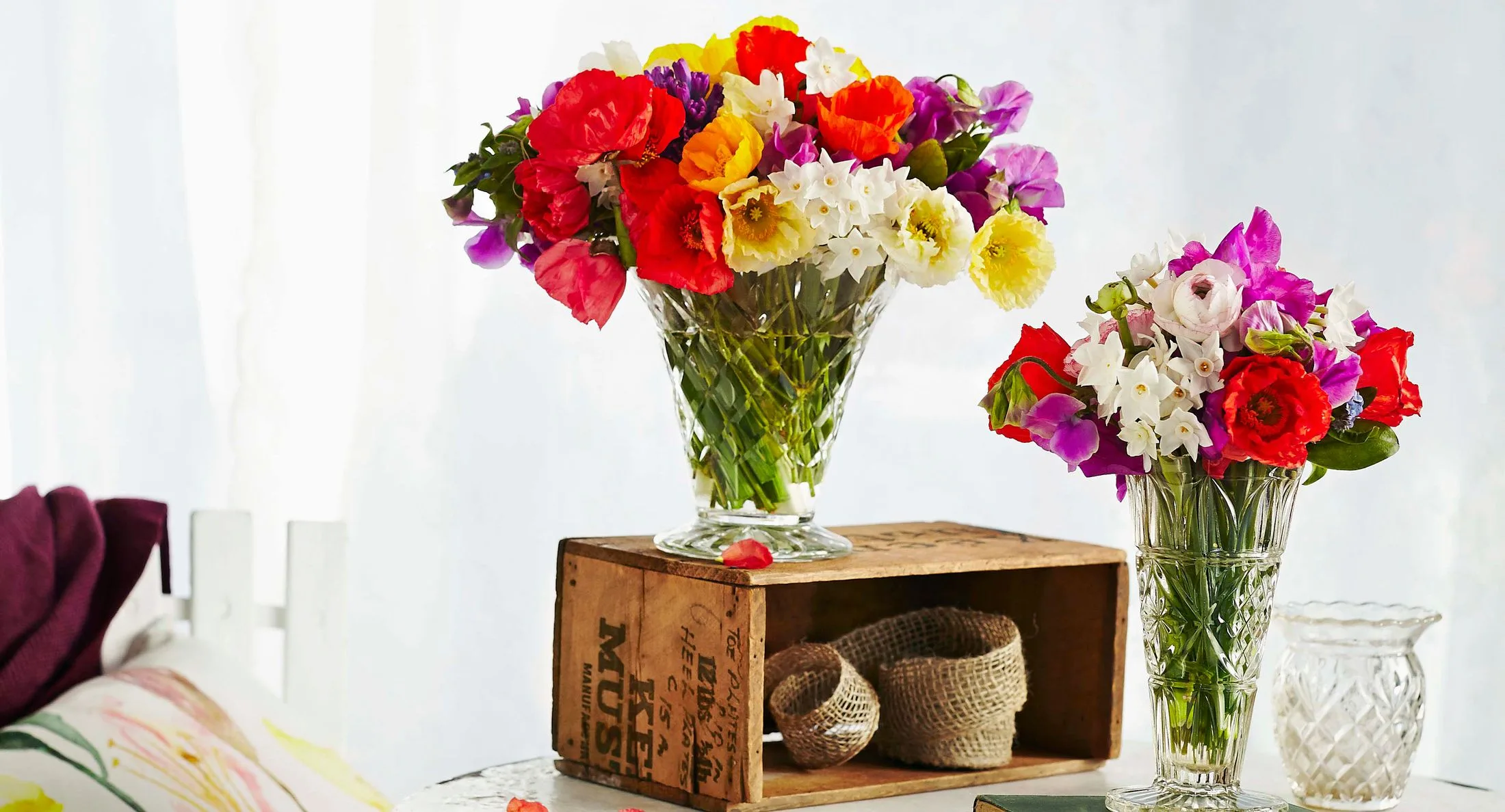 Two glass vases with vibrant mixed flowers on a table, one on a wooden crate.