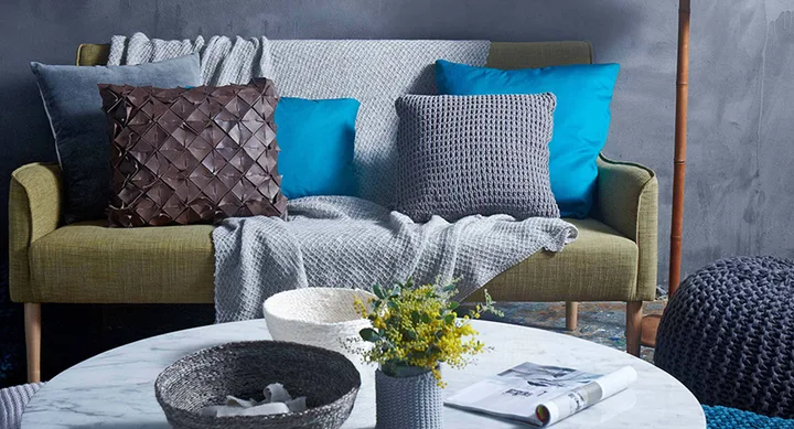 Cozy living room with a green sofa, assorted pillows, a knitted throw, and a round table with baskets and a plant.