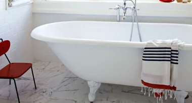Freestanding white bathtub with striped towel hanging, next to a red chair on a marble floor.