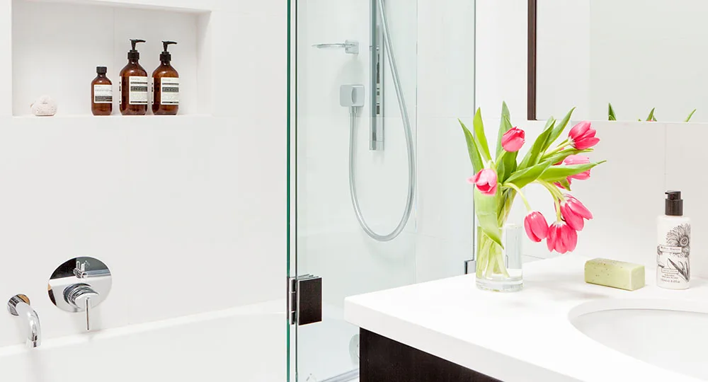 Modern white bathroom with glass shower; pink tulips in vase on counter and bottles of toiletries on shelves.