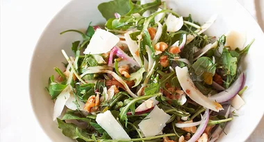 A fresh arugula salad with shaved Parmesan, red onions, walnuts, and a light vinaigrette in a white bowl.