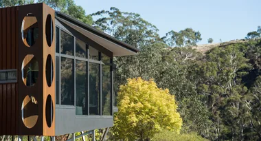Modern glass house with circular rusty panels in a wooded area.