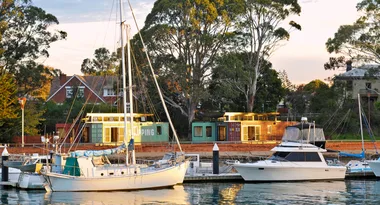 Boats docked at a marina with shipping container buildings and trees in the background during sunset.