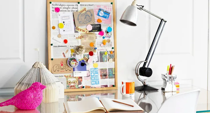 Bulletin board with colorful pins and papers, desk lamp, open notebook, pink bird figurine, and coffee mug on a desk.