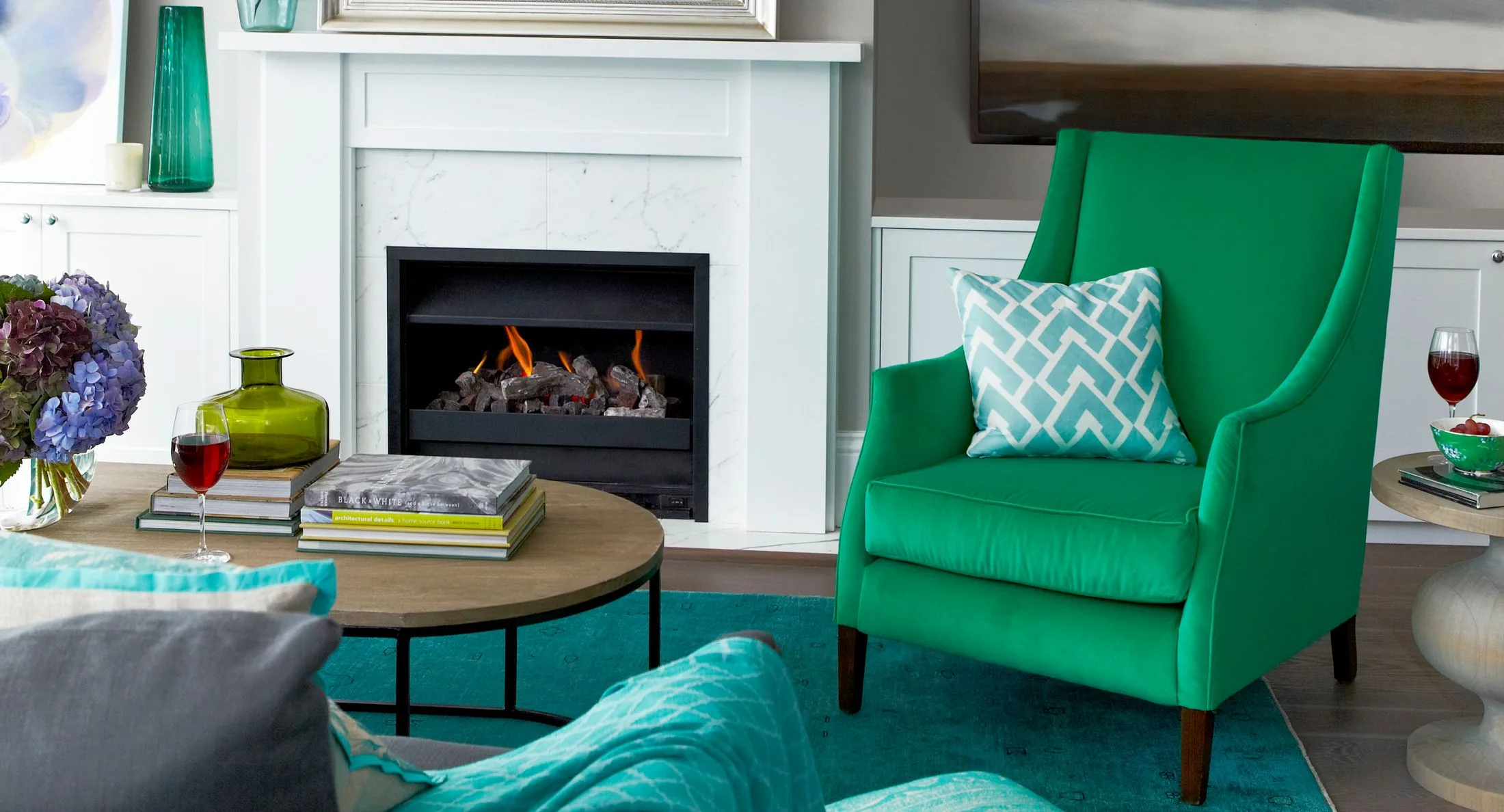 Green armchair with patterned pillow, near fireplace; coffee table with glass vase, books, and red wine glass.