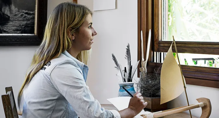 Woman with long hair writing at a desk beside a window, with feathers and a small sailboat in view.