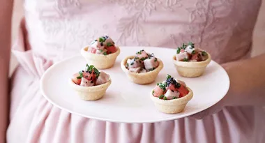 A person in a pink dress holding a plate with five small appetizer tarts topped with diced ingredients and herbs.