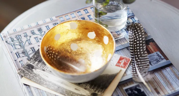 Golden decorative bowl with white spots, placed on a book and photo, alongside a feather and a glass of water.