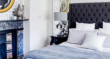 Elegant bedroom with dark tufted headboard, decorative lamp, mirrored wall, and ornate fireplace with floral tiles.