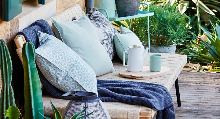 Outdoor lounge with cushions, cactus plant, blue blanket, white teapot, and green mug on a wooden surface.
