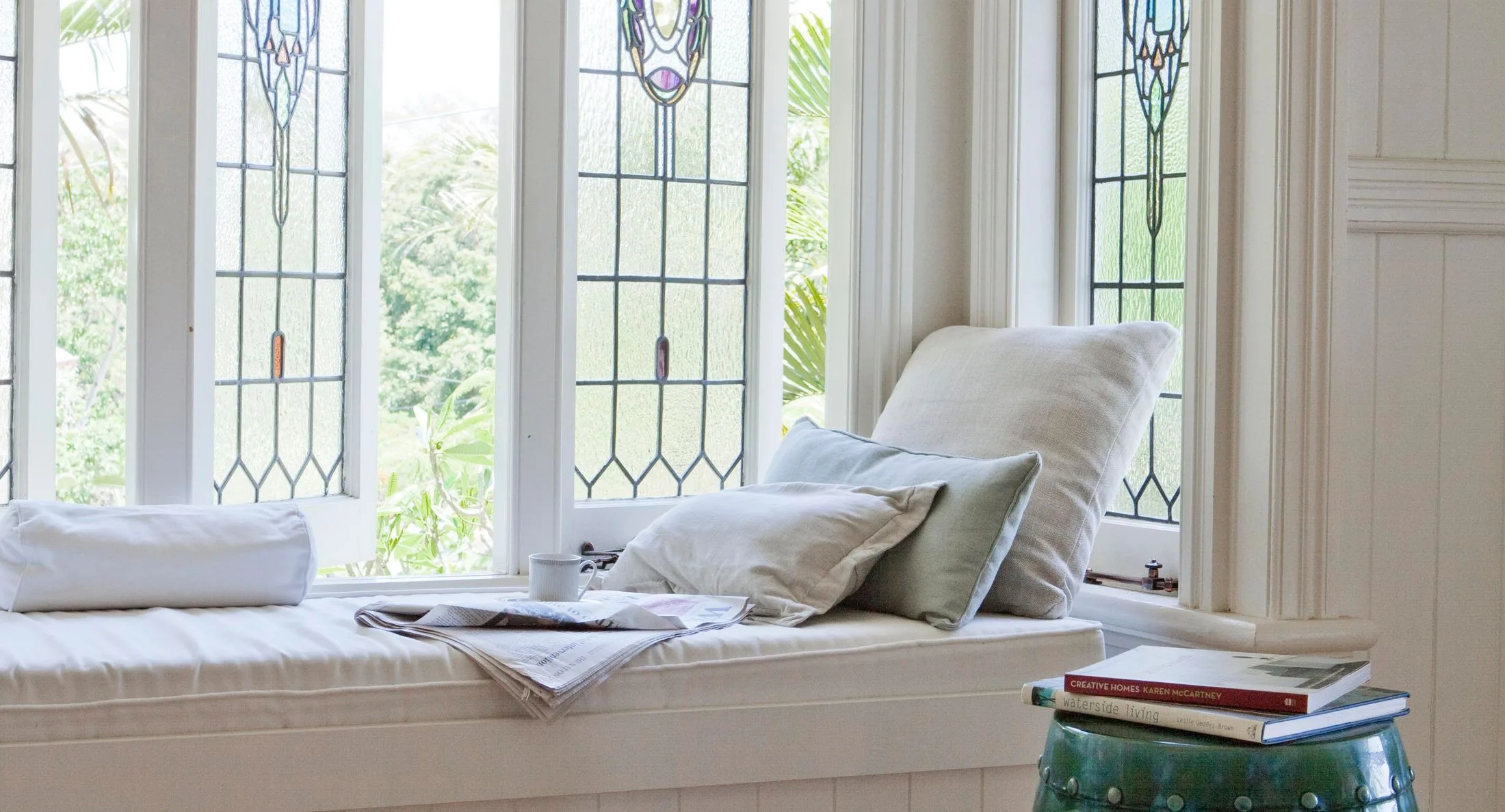 Cozy window seat with cushions, a cup, and books, next to stained glass windows and greenery outside.
