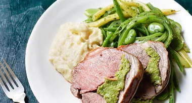 Sliced beef with green sauce, mashed potatoes, and mixed vegetables on a white plate with a fork beside it.