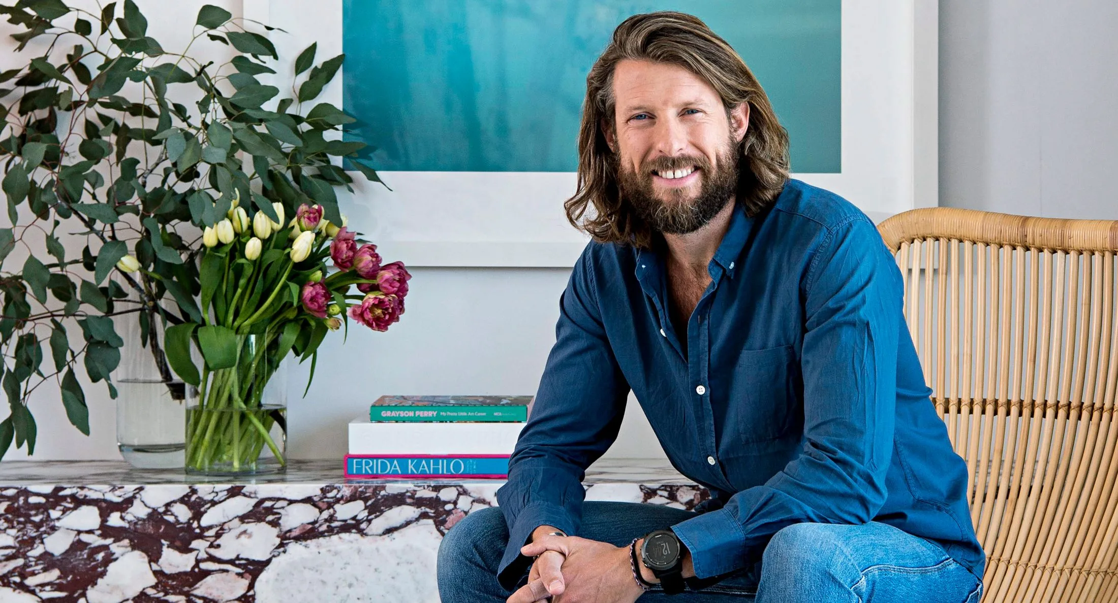 A man with long hair and beard sitting on a chair by a table with flowers and books, smiling at the camera.