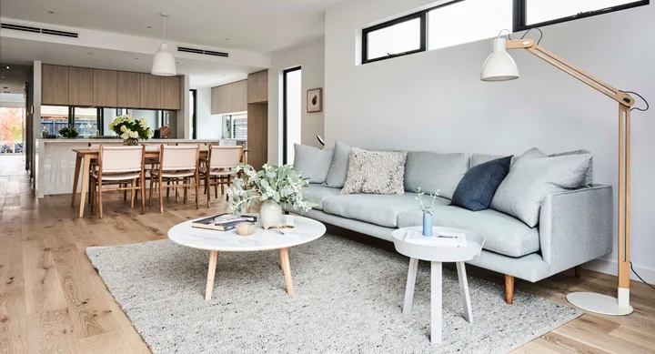 Modern living room with a light gray sofa, wooden floor, round coffee table, and dining area in the background.