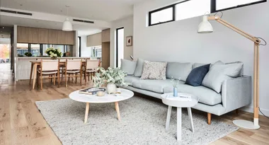 Modern living room with a light gray sofa, wooden floor, round coffee table, and dining area in the background.