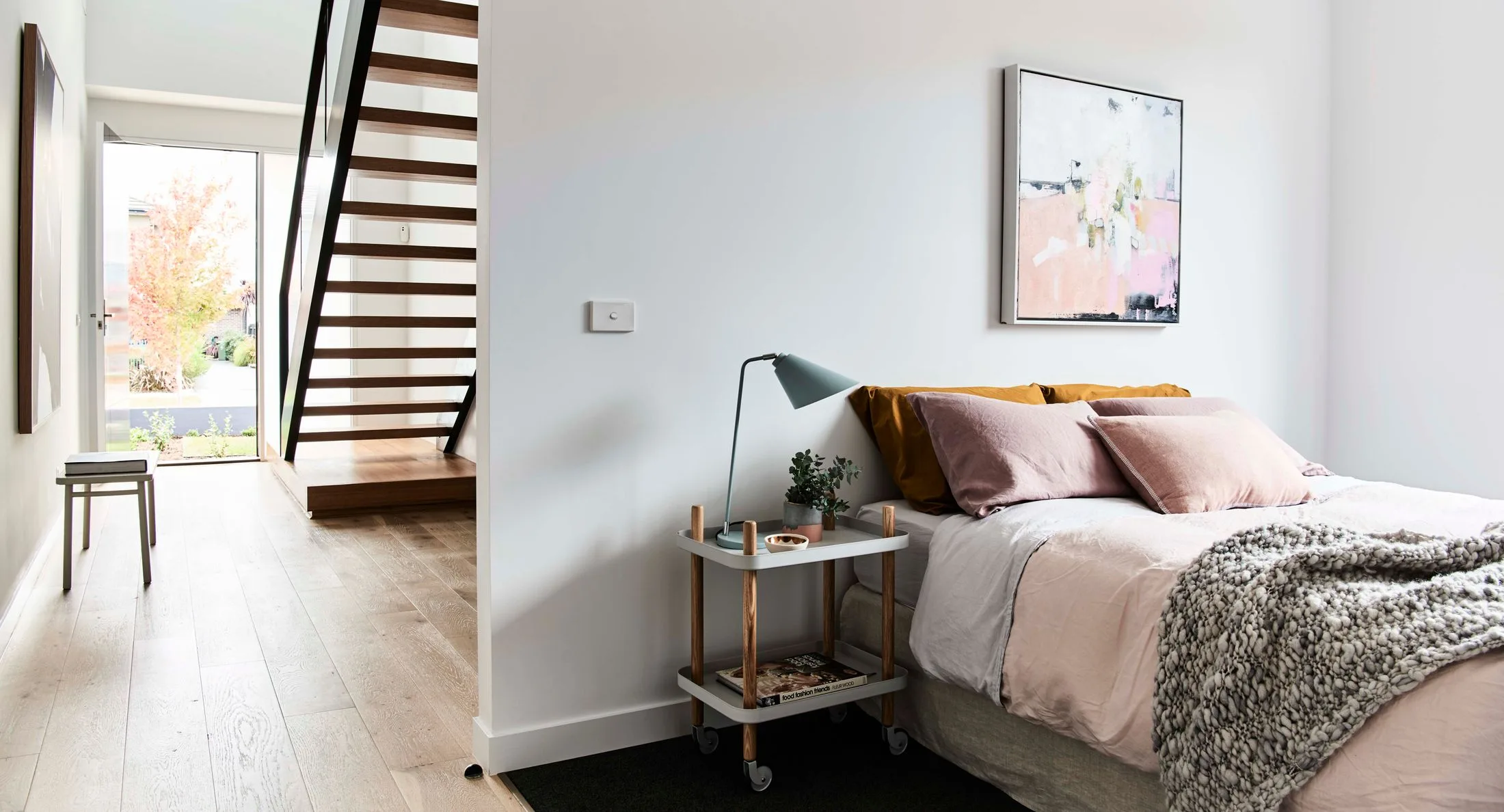 Bedroom with a wooden staircase, a bed with pastel pillows, a side table with a lamp, and abstract wall art.