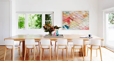 Bright dining room with a wooden table, white chairs, colorful artwork, and a vase of flowers by the window.