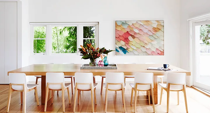 Bright dining room with a wooden table, white chairs, colorful artwork, and a vase of flowers by the window.
