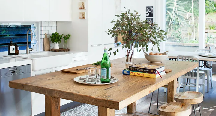 Rustic kitchen with wooden table, vase with branches, books, and a green bottle; modern white cabinetry in background.