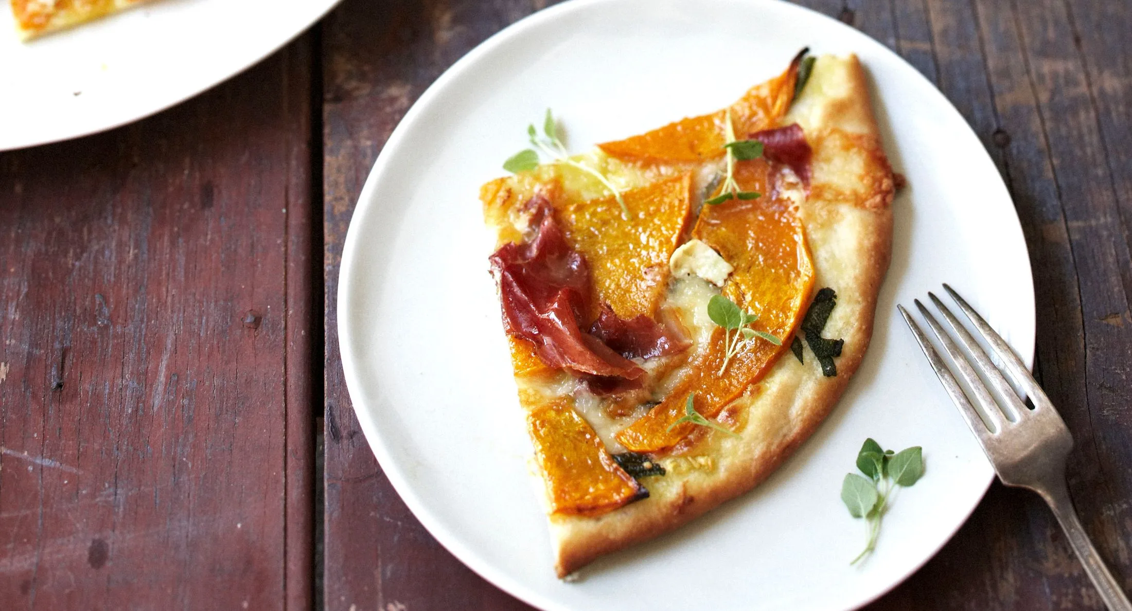 Slice of gourmet pizza with prosciutto, roasted squash, and fresh herbs on a white plate.