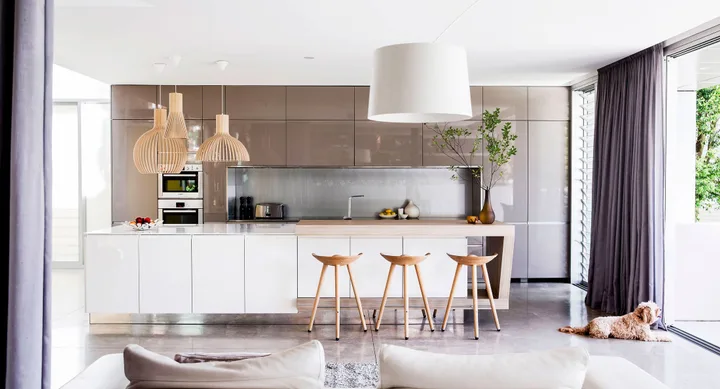 Modern kitchen with beige cabinets, white island, wooden stools, pendant lights, and a dog by the window.
