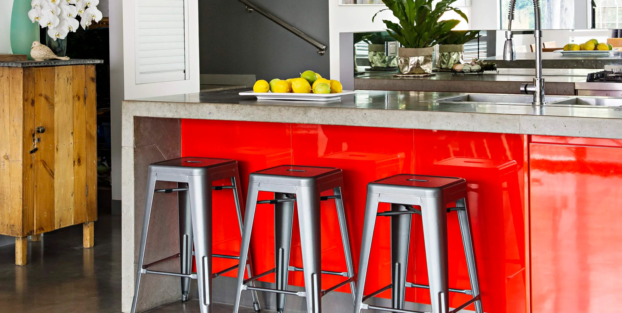 Modern kitchen with red cabinets, concrete countertop, metal bar stools, and a fruit bowl.