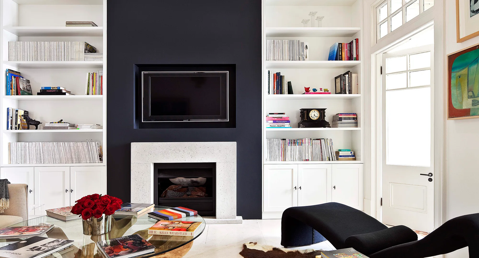 Living room with a TV above a white fireplace, surrounded by shelves of books and magazines, and a vase of red roses on the table.