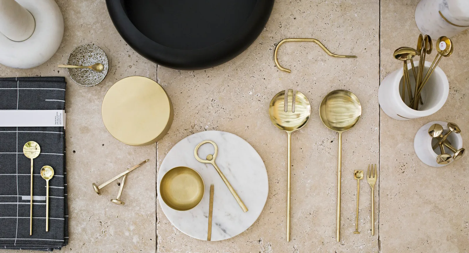 Gold kitchen utensils on a stone surface with a black plate, marble dish, and gray napkin.