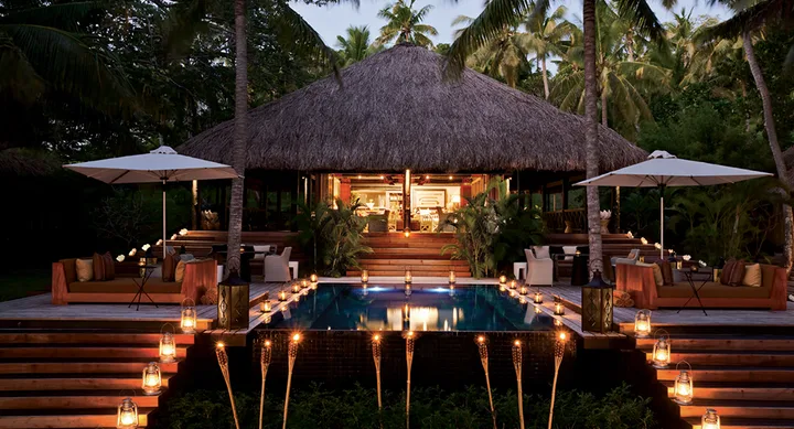 Tropical villa at night with pool, tiki torches, and ambient lighting, surrounded by palm trees.