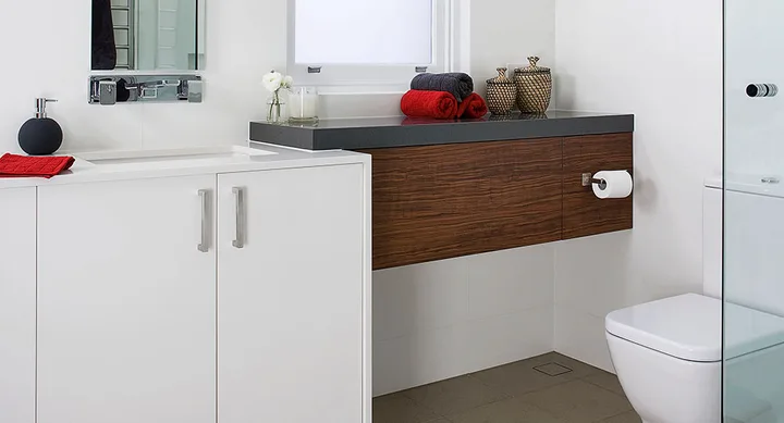 Modern bathroom with white and wood cabinetry, red and black towels, glass shower, and toilet.