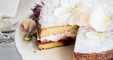 Victoria sponge cake dusted with sugar, layered with cream and strawberries, topped with rose petals on a cake stand.