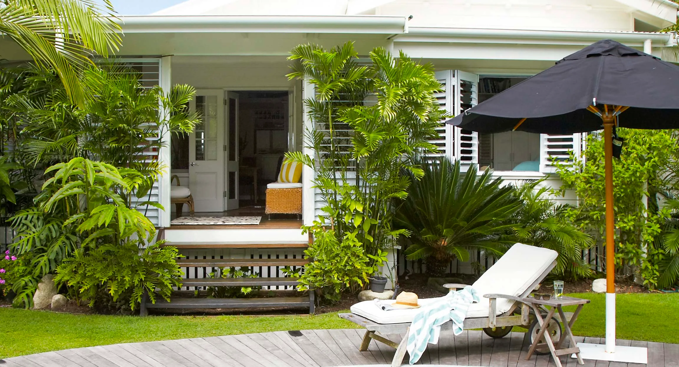 Outdoor patio with lush plants, a lounge chair, table, and large black umbrella in front of a white house.