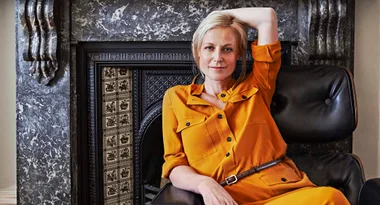 A woman in an orange outfit sits on a black chair, posing with one arm behind her head in front of a decorative fireplace.