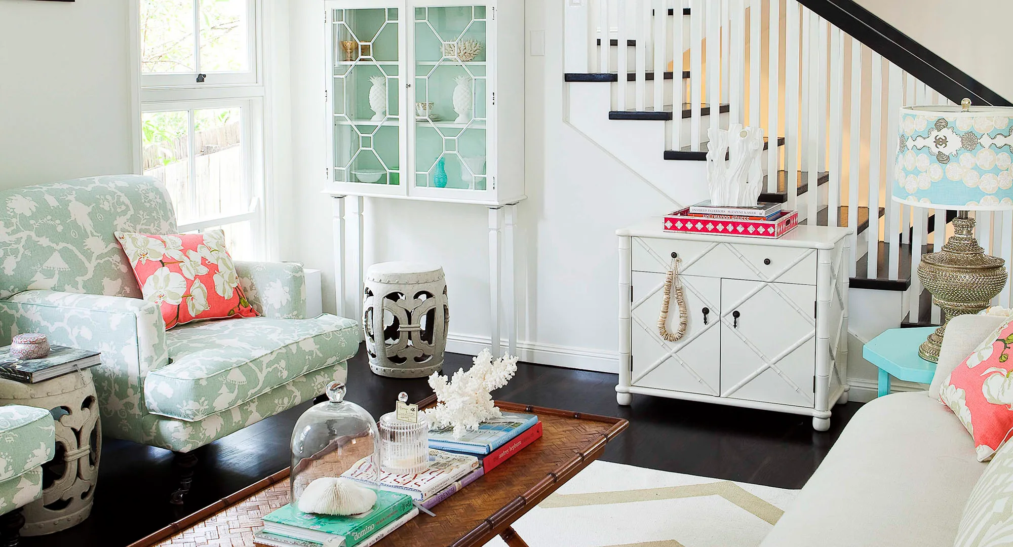 Cozy living room with floral armchair, decorative pillows, books on coffee table, and white cabinet under staircase.