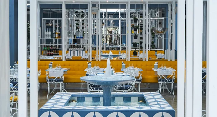 Restaurant interior with blue and white decor, patterned fountain, yellow seating, and white metal chairs.