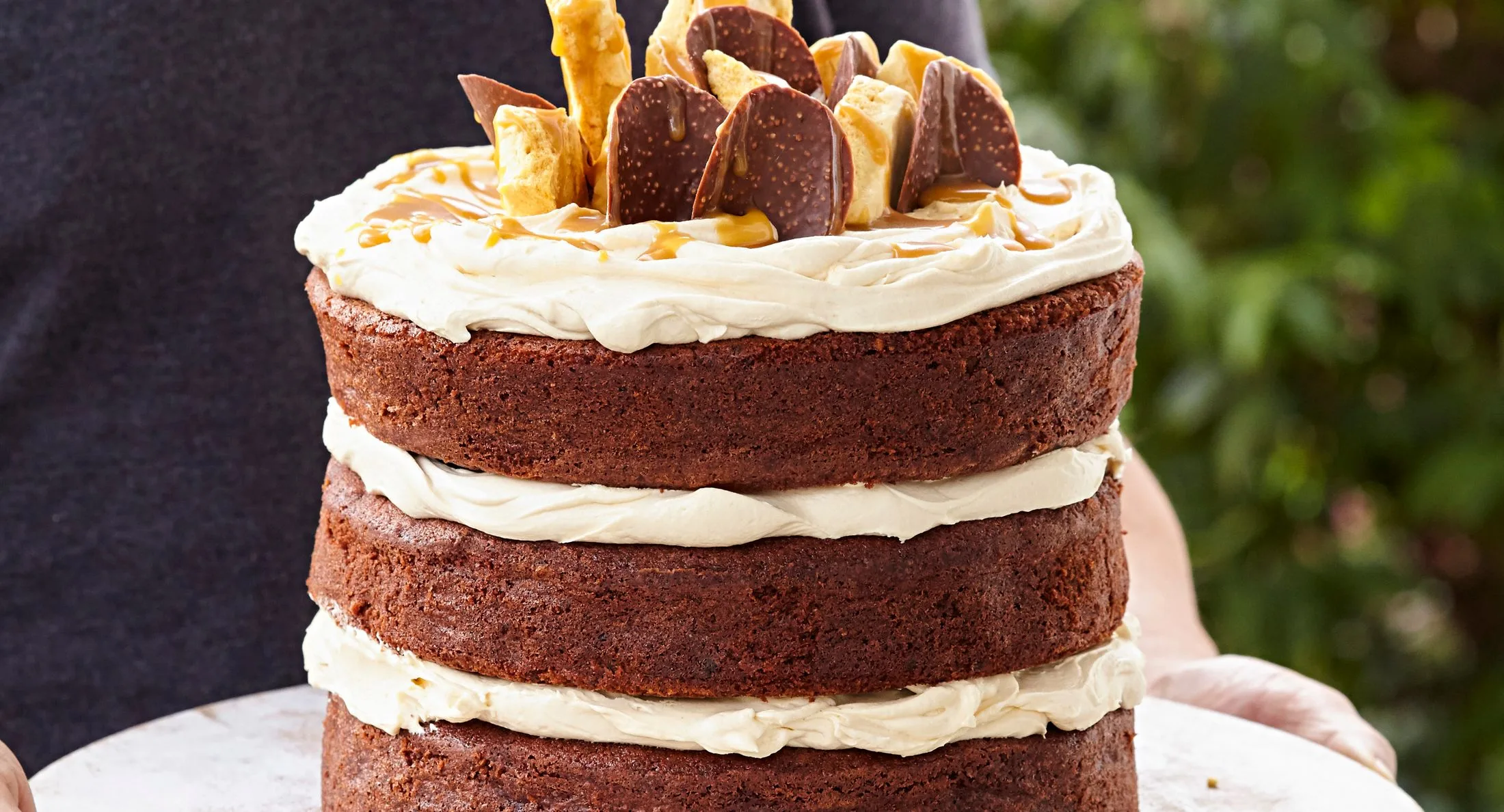 Three-layer chocolate cake with cream frosting, topped with chocolate pieces and honeycomb, held by a person against greenery.