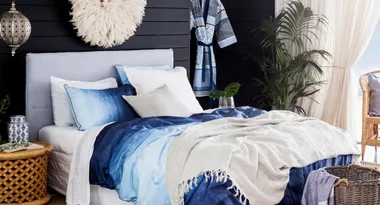 Cozy bedroom with blue and white bedding, plant decor, wicker basket, and a hanging robe beside a window.
