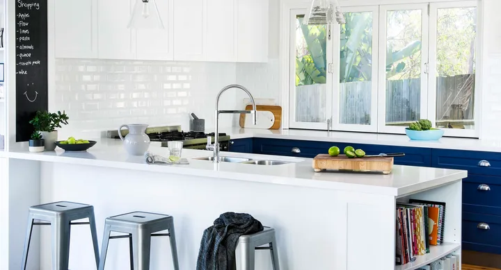 Modern kitchen with white cabinets, blue drawers, island, stools, and chalkboard list, under natural light.