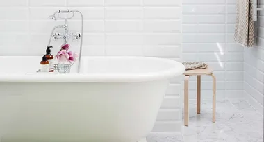 Freestanding bathtub with chrome faucet, flowers, soap bottle, and stool in a white-tiled bathroom.