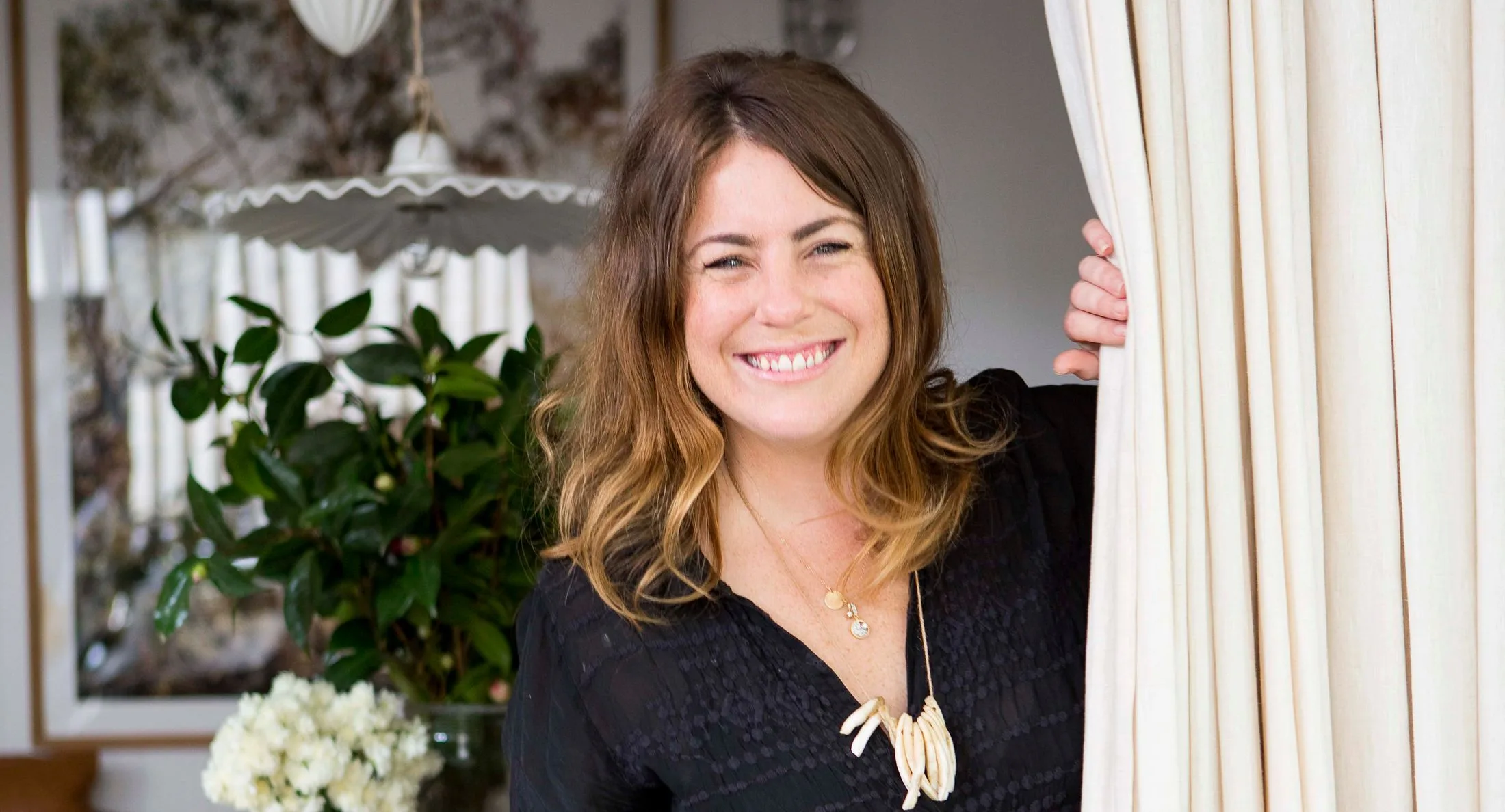 Smiling woman peeking from behind a curtain in a room with plants and decorative lighting.