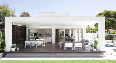 Outdoor patio with modern white furniture, dining and seating areas, surrounded by greenery and a pool in the background.
