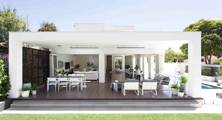 Outdoor patio with modern white furniture, dining and seating areas, surrounded by greenery and a pool in the background.