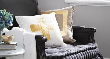 Alt text: "Armchair with gold-accented pillows, gray blanket, and side table with vase and gold bowl."