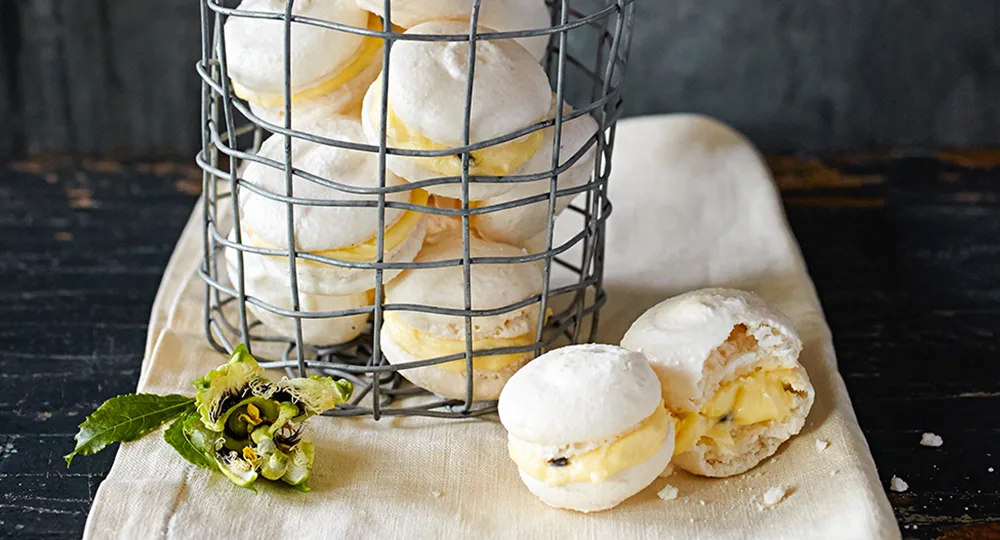 White macarons with yellow filling in a wire basket, on a cloth, with a passionfruit flower beside.