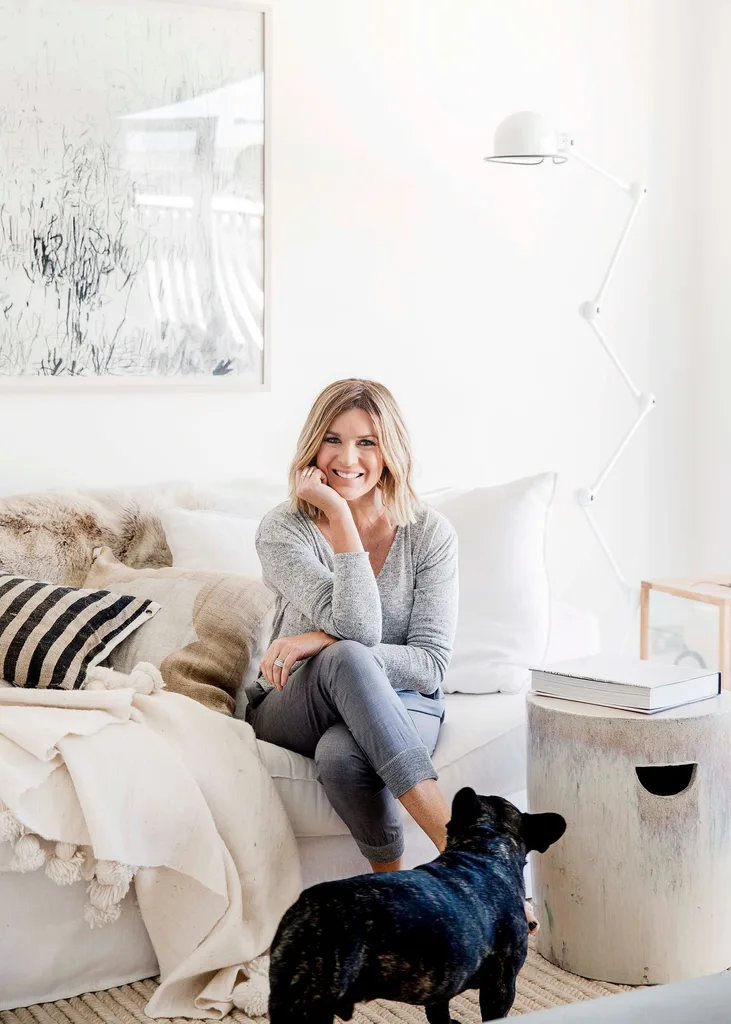 A woman sitting on a white sofa with a black dog nearby in a bright living room.